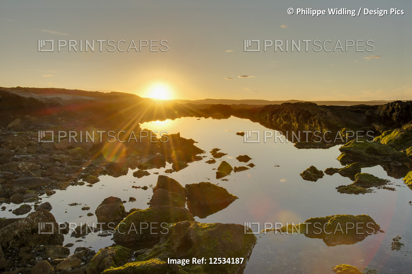 A golden-coloured sunset and sunburst hilights the rocks around an ocean-side ...