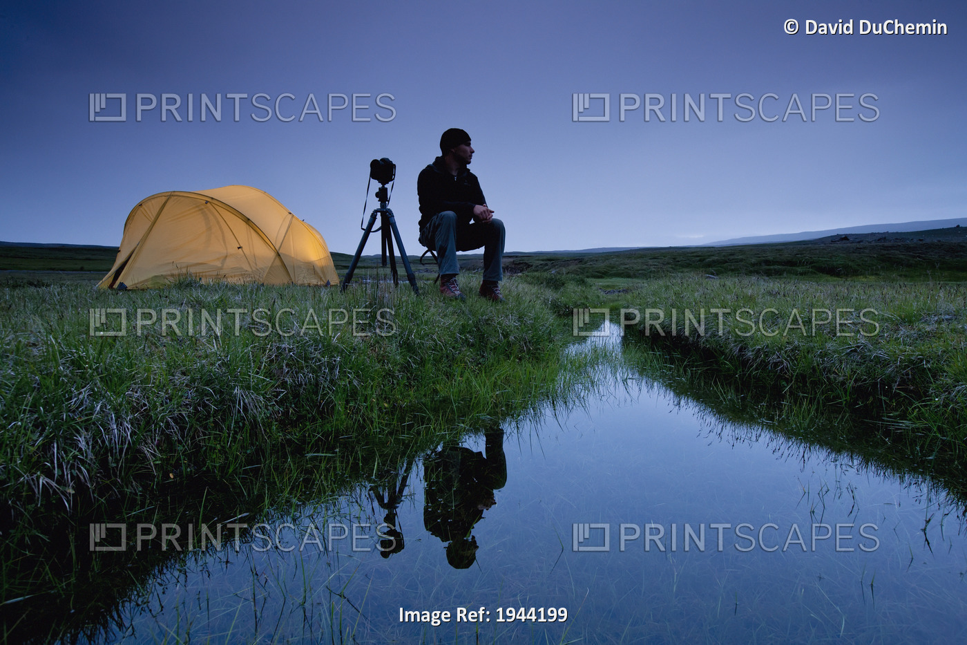 A Photographer Sits By His Tent And A Stream Of Water; Iceland