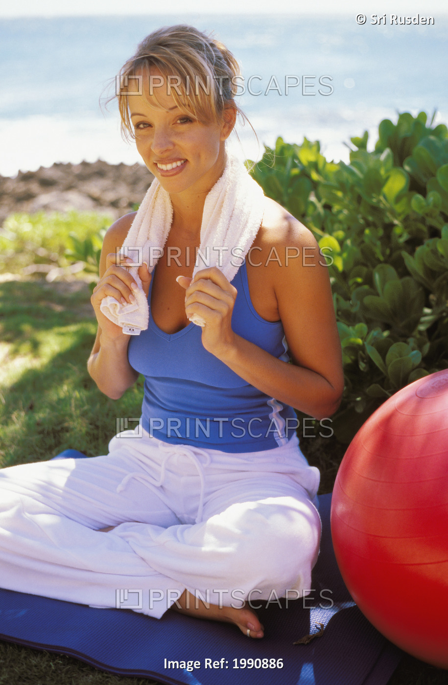 Woman Sitting In Yoga Position Near Ocean On Mat With Red Exercise Ball