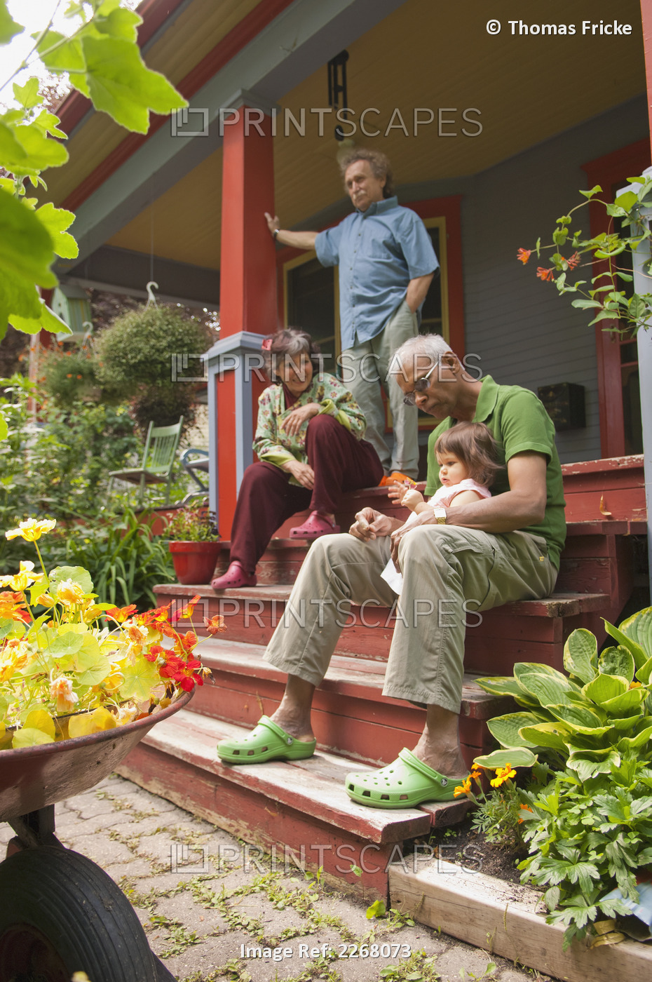 Family Sitting On Front Porch Of Home Surrounded By Plants And Flowers; ...
