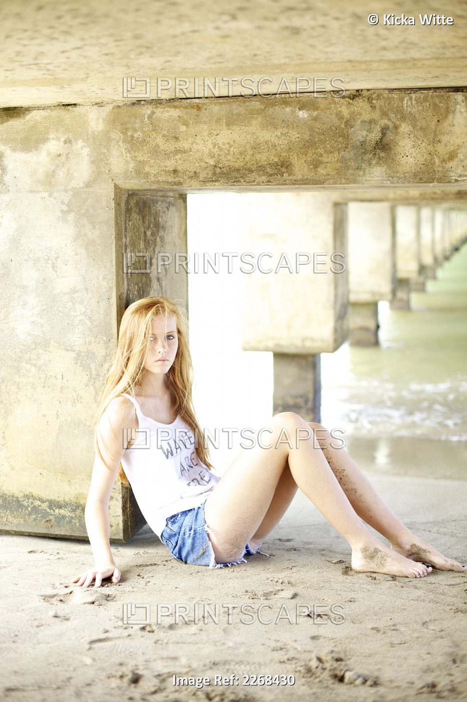 A young woman with long red hair sits in the sand under a bridge at the water's ...
