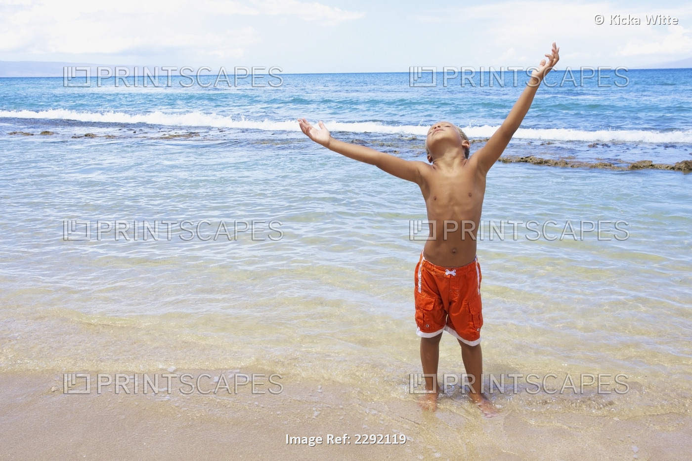 A young boy standing in the shallow water of the ocean with arms raised and ...