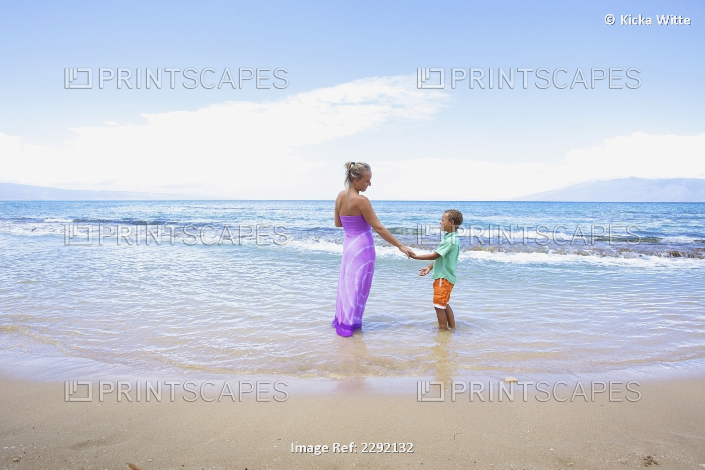 A mother and son hold hands while standing in the shallow water at the ocean; ...