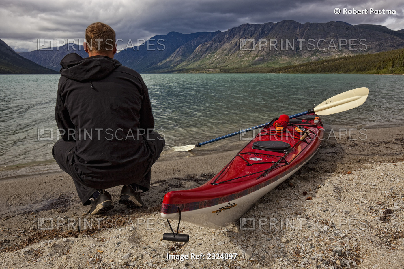 Kayaker Squatting By His Kayak On The Shoreline Of Kusawa Lake; Yukon, Canada