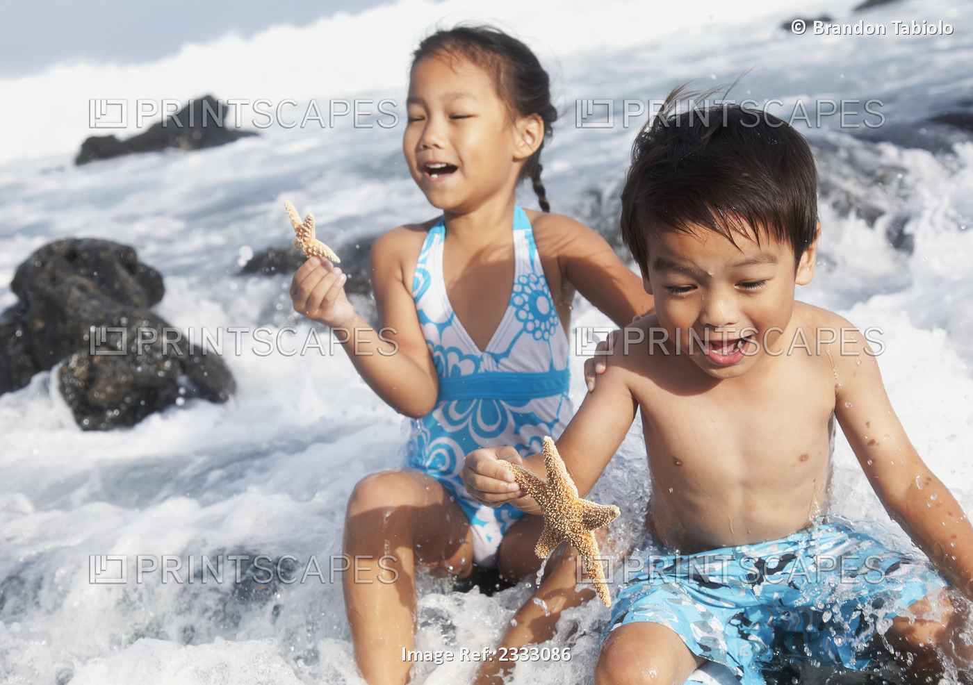 A young boy and girl hold starfish and sit in the crashing waves at the water's ...