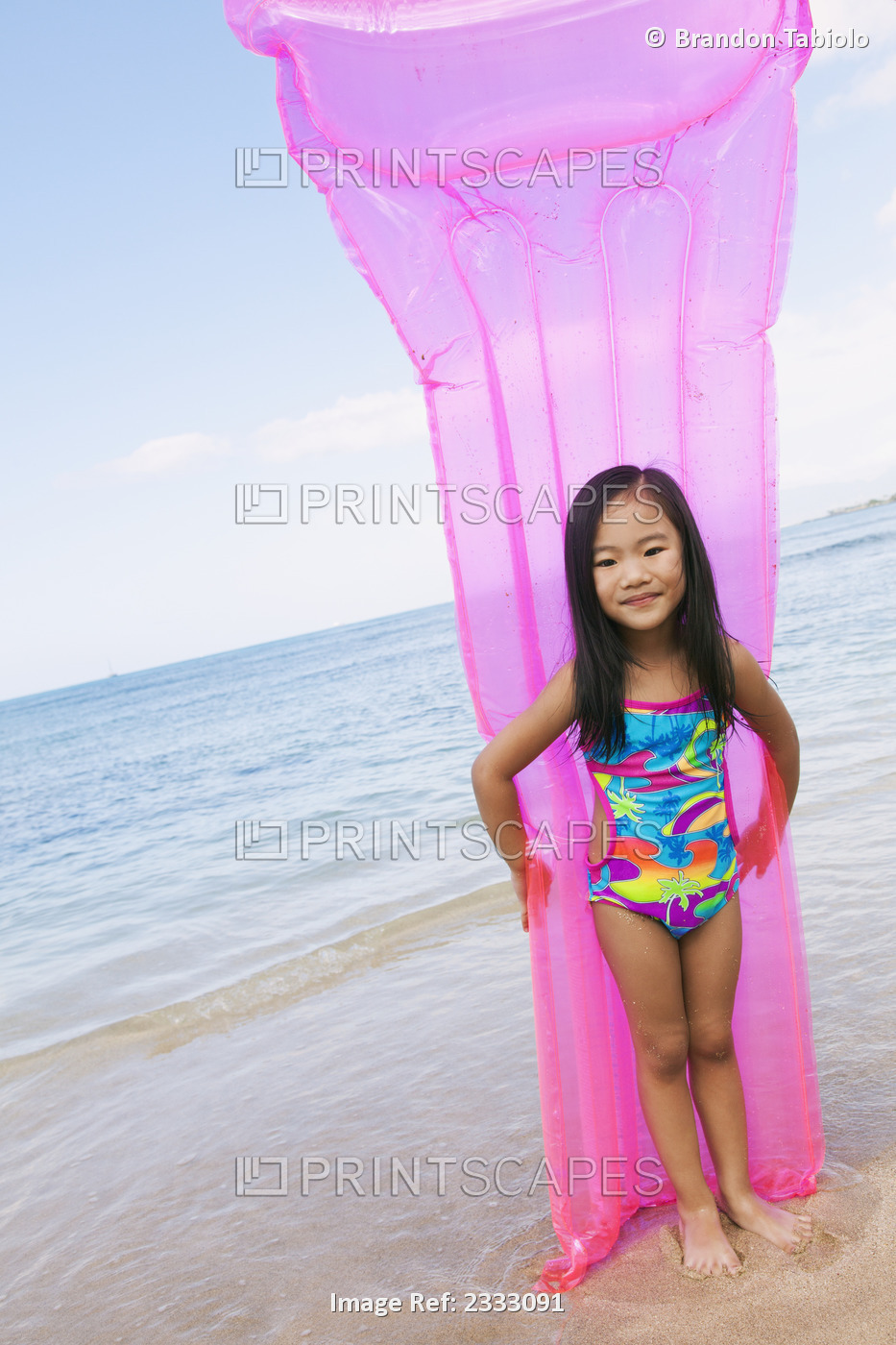 A young girl in a swimsuit holding an inflatable mattress at the water's edge; ...