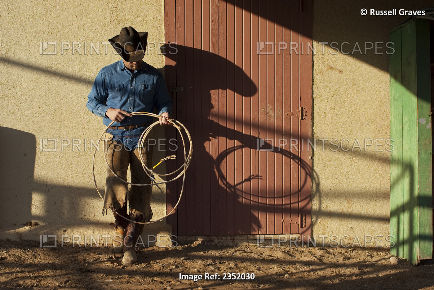 Agriculture - Cowboy with a lasso rope standing against a barn in evening light ...
