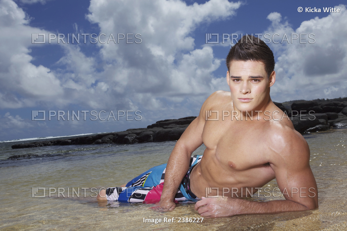 A Young Man In A Bathing Suit Lays In The Shallow Ocean Water At The Beach; ...