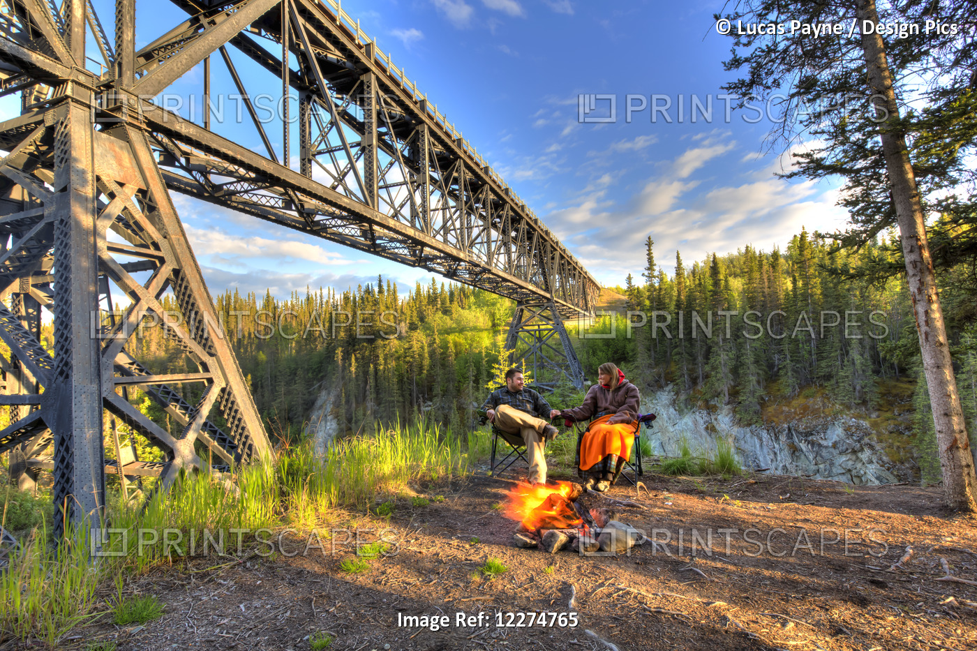 Couple Sitting By A Campfire Holding Hands Under The Kuskulana River Bridge ...