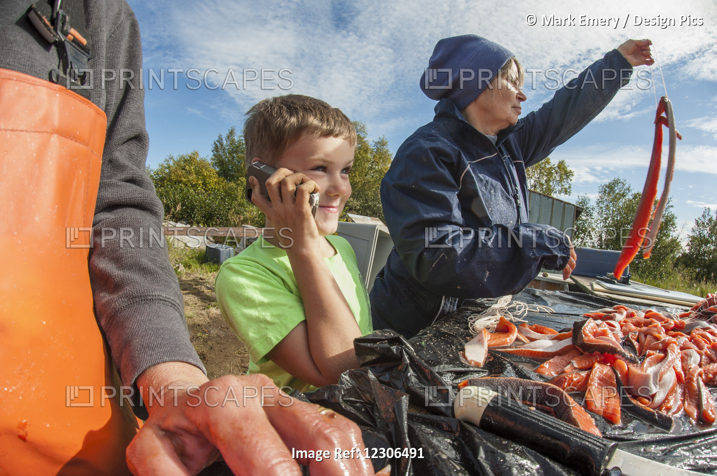 Young Boy Talks On A Cell Phone While Adults Filet And Hang Caught Salmon, ...