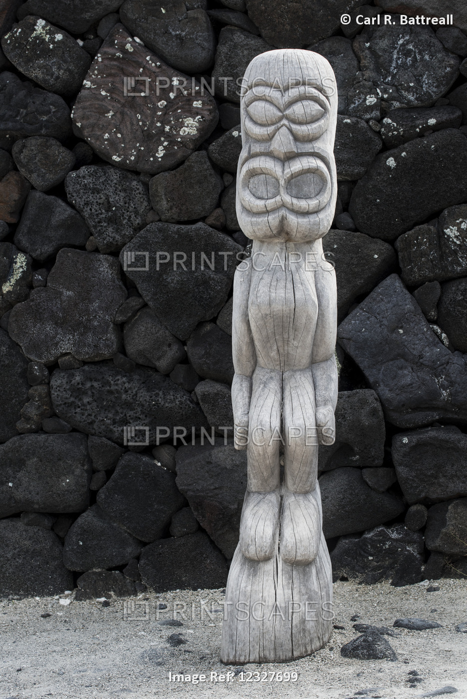 Carved Wooden Sculpture Standing Against A Wall Of Black Rock; Hawaii, United ...