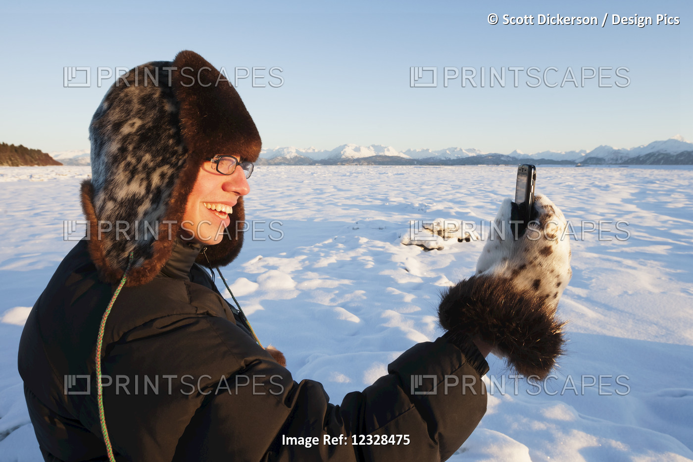 Man standing in snow with a fur hat and mittens using a cell phone, ...