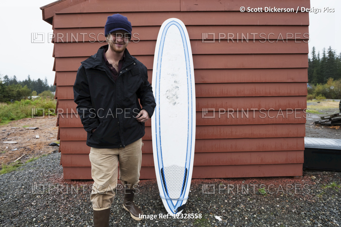 Young man standing next to a surfboard, South-central Alaska; Homer, Alaska, ...