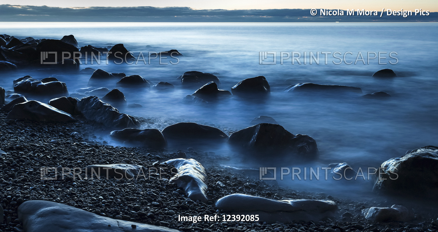 Beautiful blue misty ocean washing in against rocks on a West Coast beach; ...