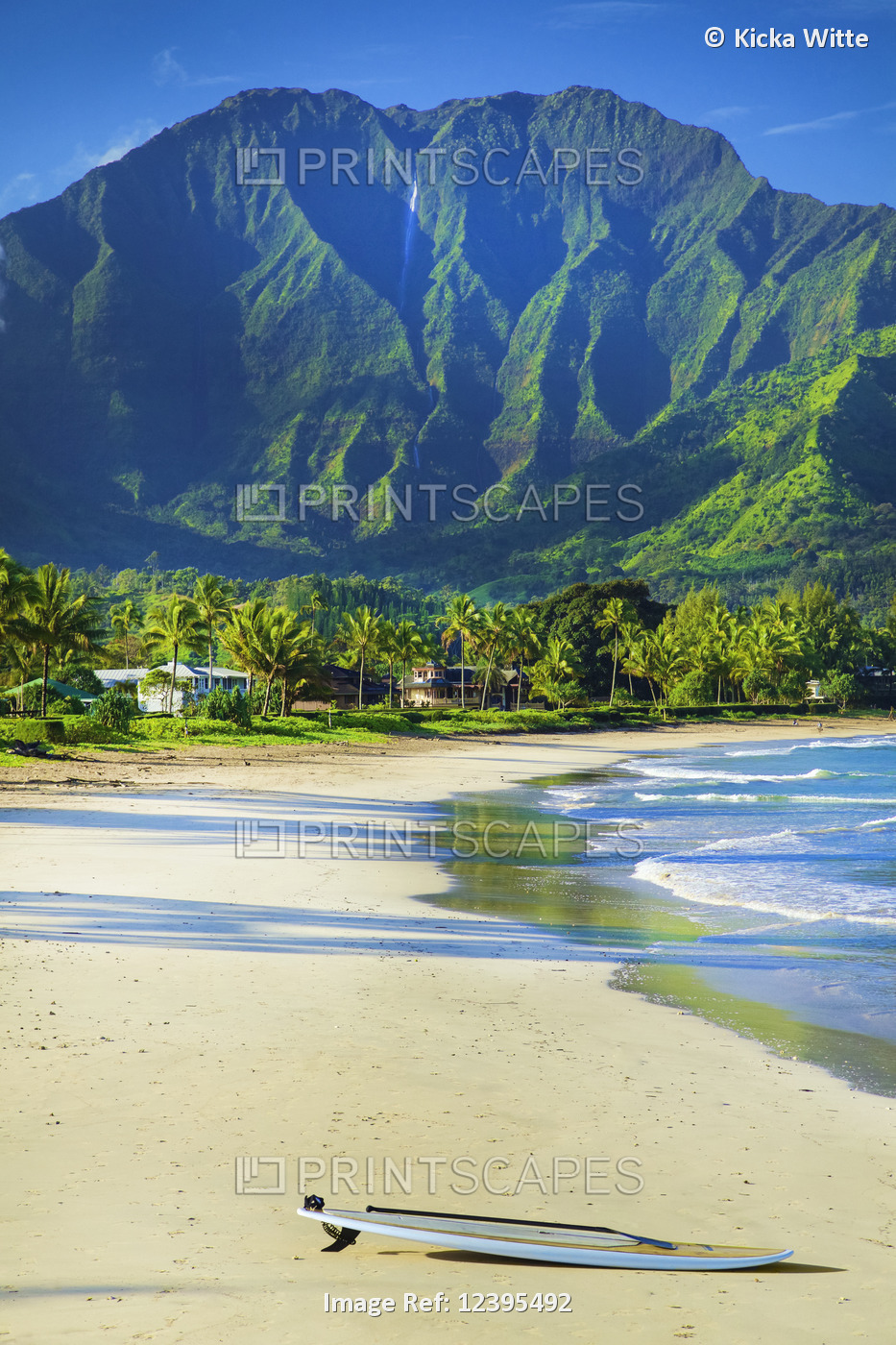A surfboard sits on the beach at the water's edge with rugged green mountains ...