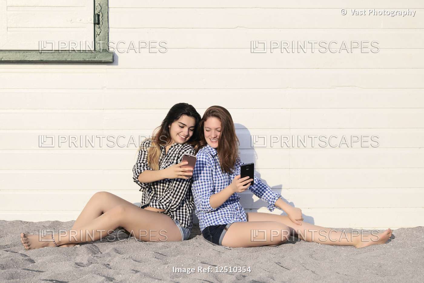 Two teenage girls sitting in the sand against a building looking at their smart ...