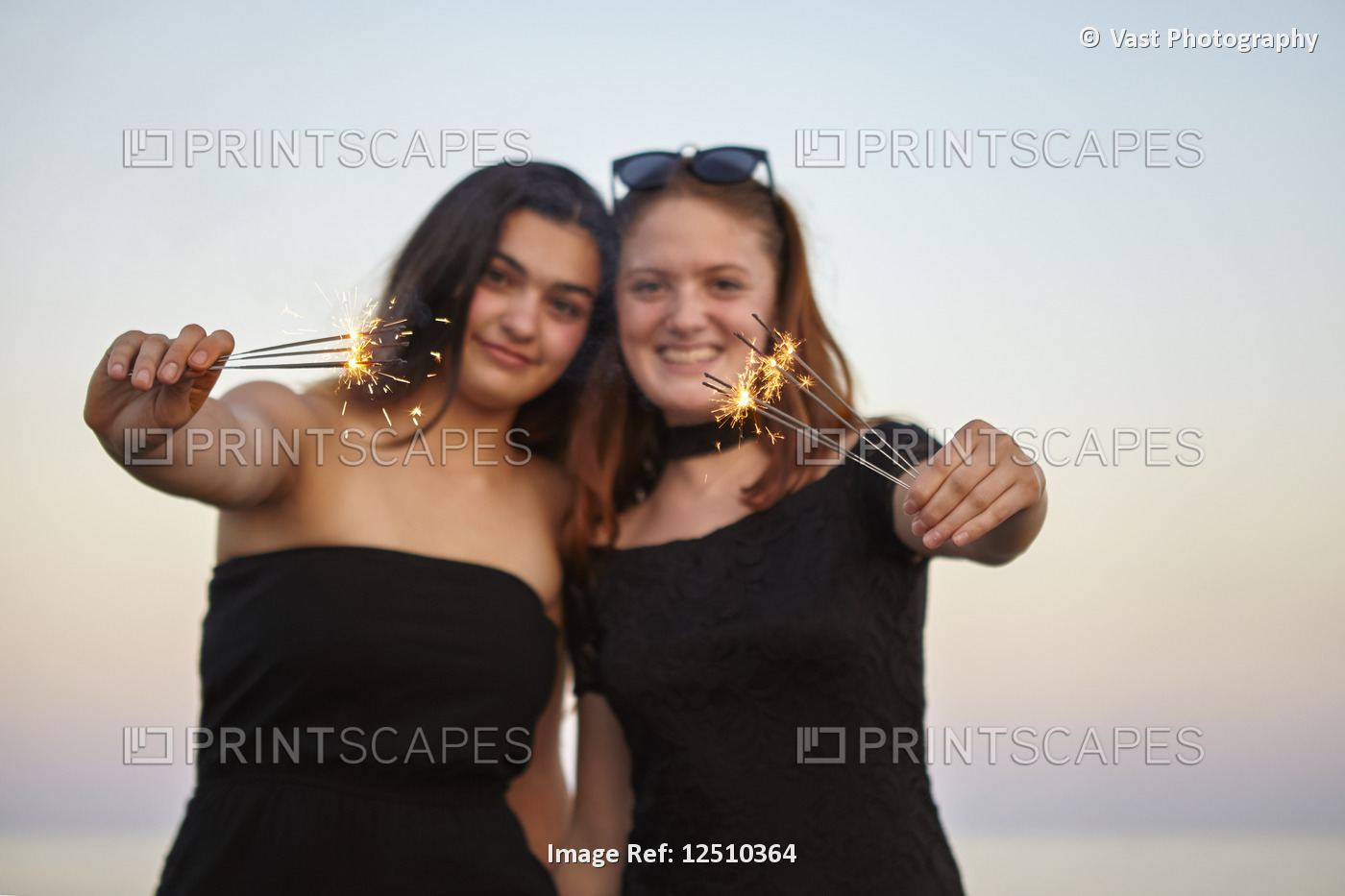 Two teenage girls in formal black dresses hold out sparklers towards the camera ...