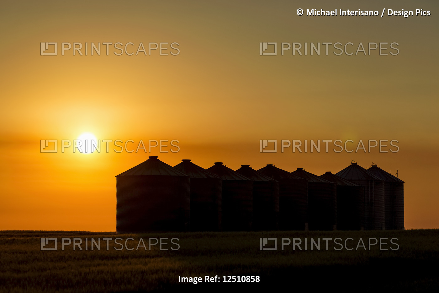 Silhouette of large metal grain bins at sunrise with an orange sun and an ...