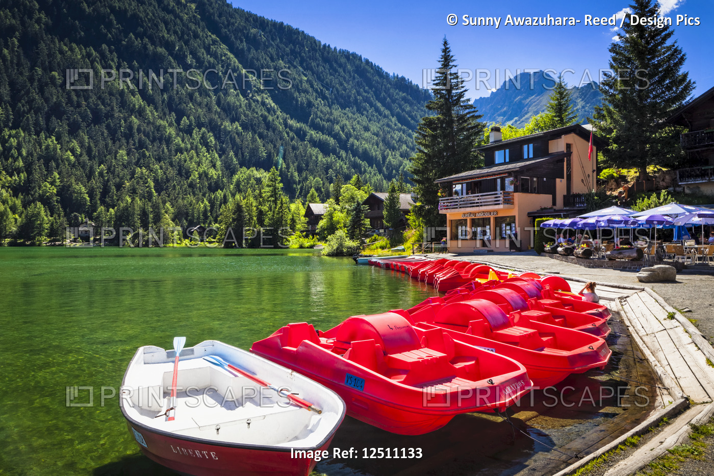 Red boats lined up at Champex Lake with an outdoor cafe under blue sky; ...