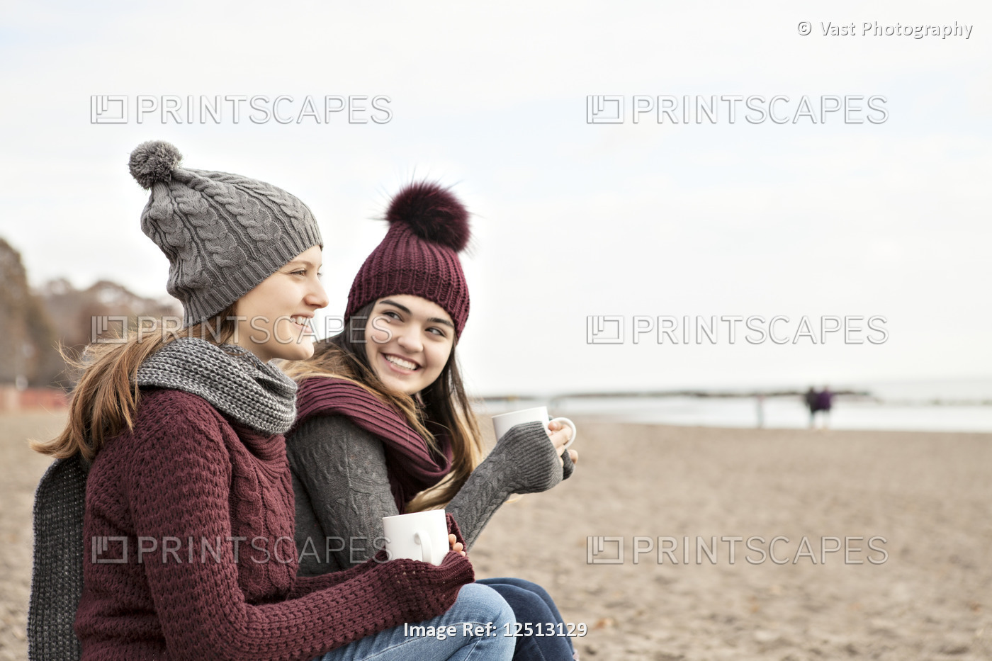 Two friends at the beach wearing knit hats and scarves, sitting and having ...