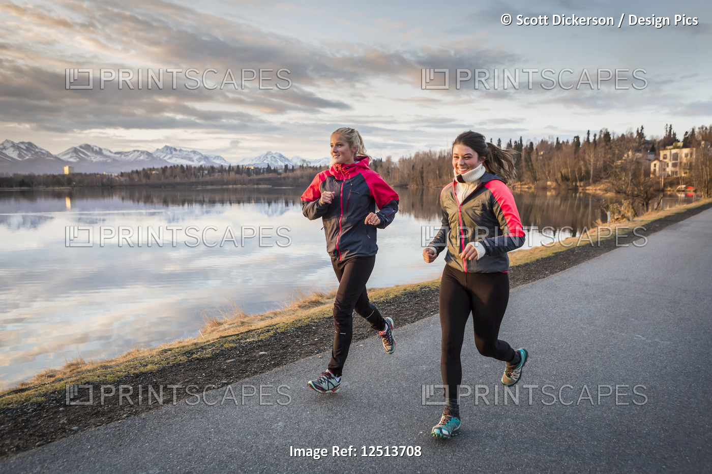 Two young women running on a trail at the water's edge with mountains in the ...