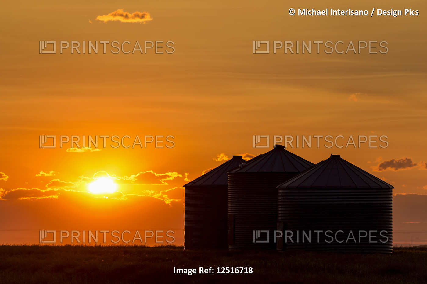 Silhouette of large metal grain bins at sunrise with orange sun rising over ...