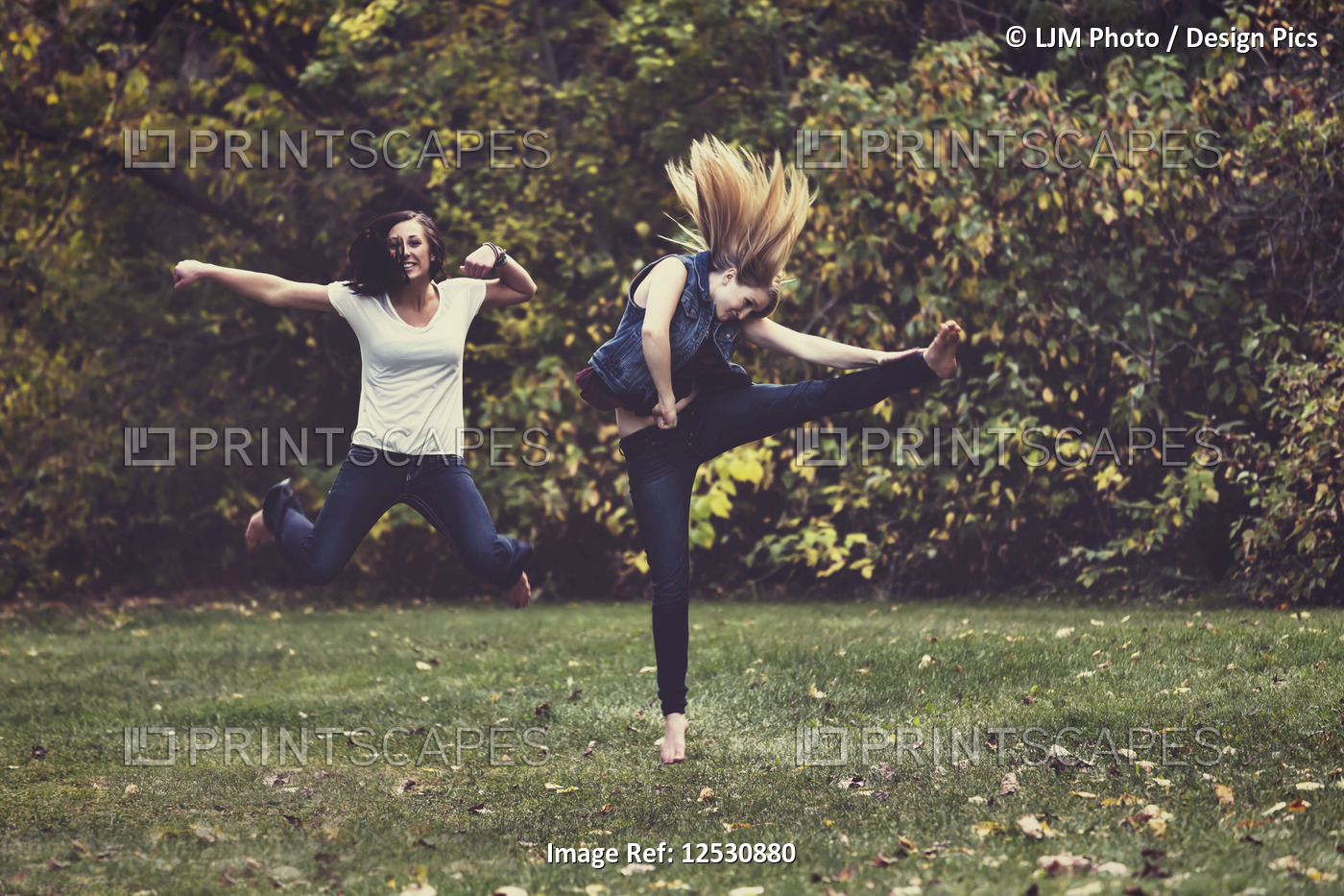 Two young girls kicking up their heels and leaping and dancing in a park in the ...