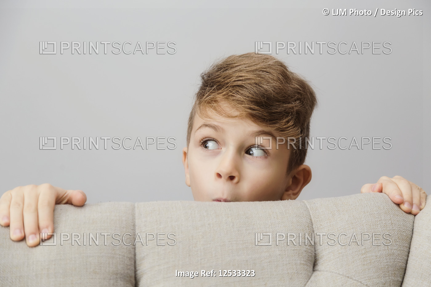 Portrait of a young boy peeking out from behind a couch with a silly facial ...