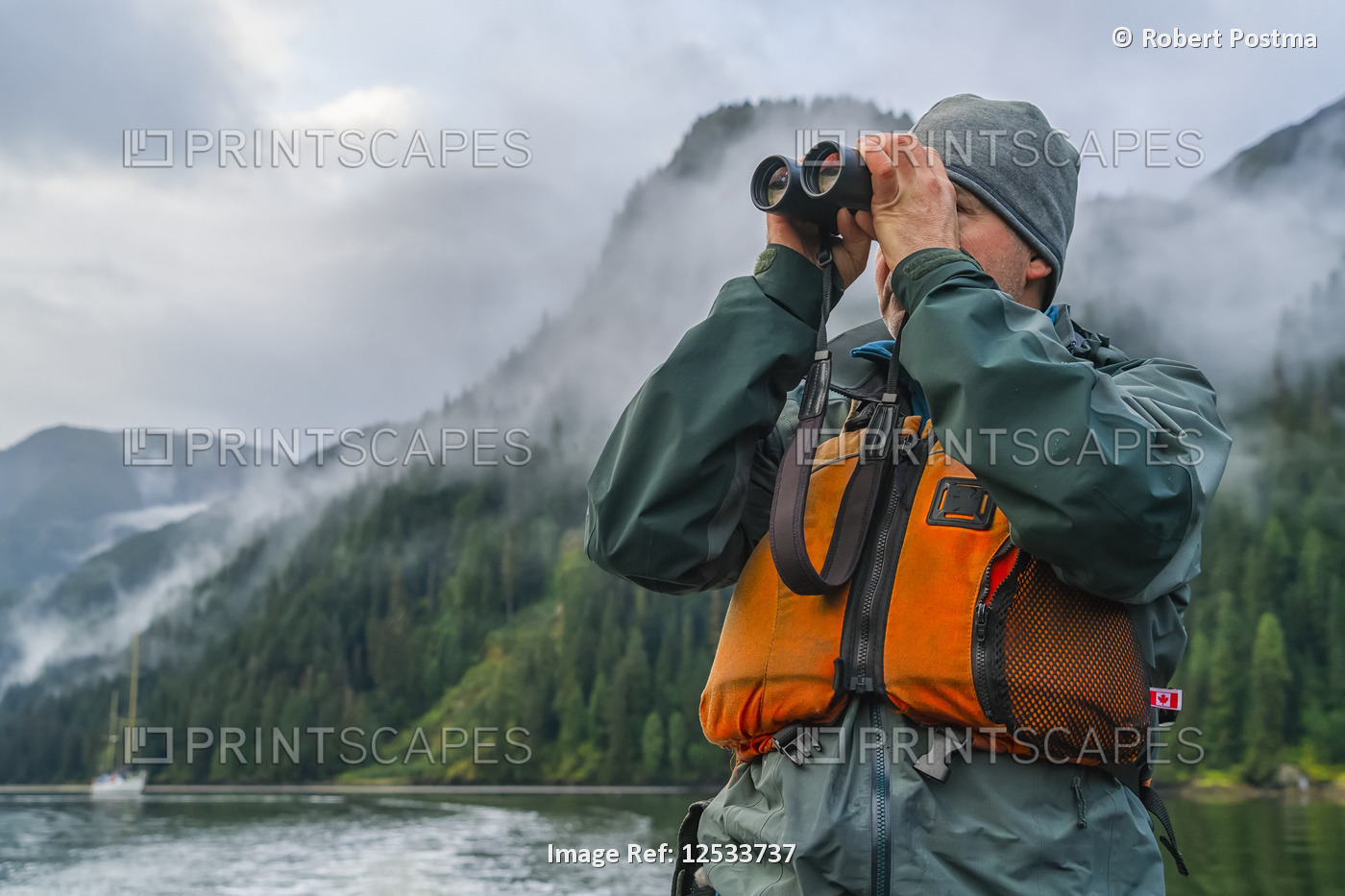 Man looking through a pair of binoculars with his sailboat in the distance, ...