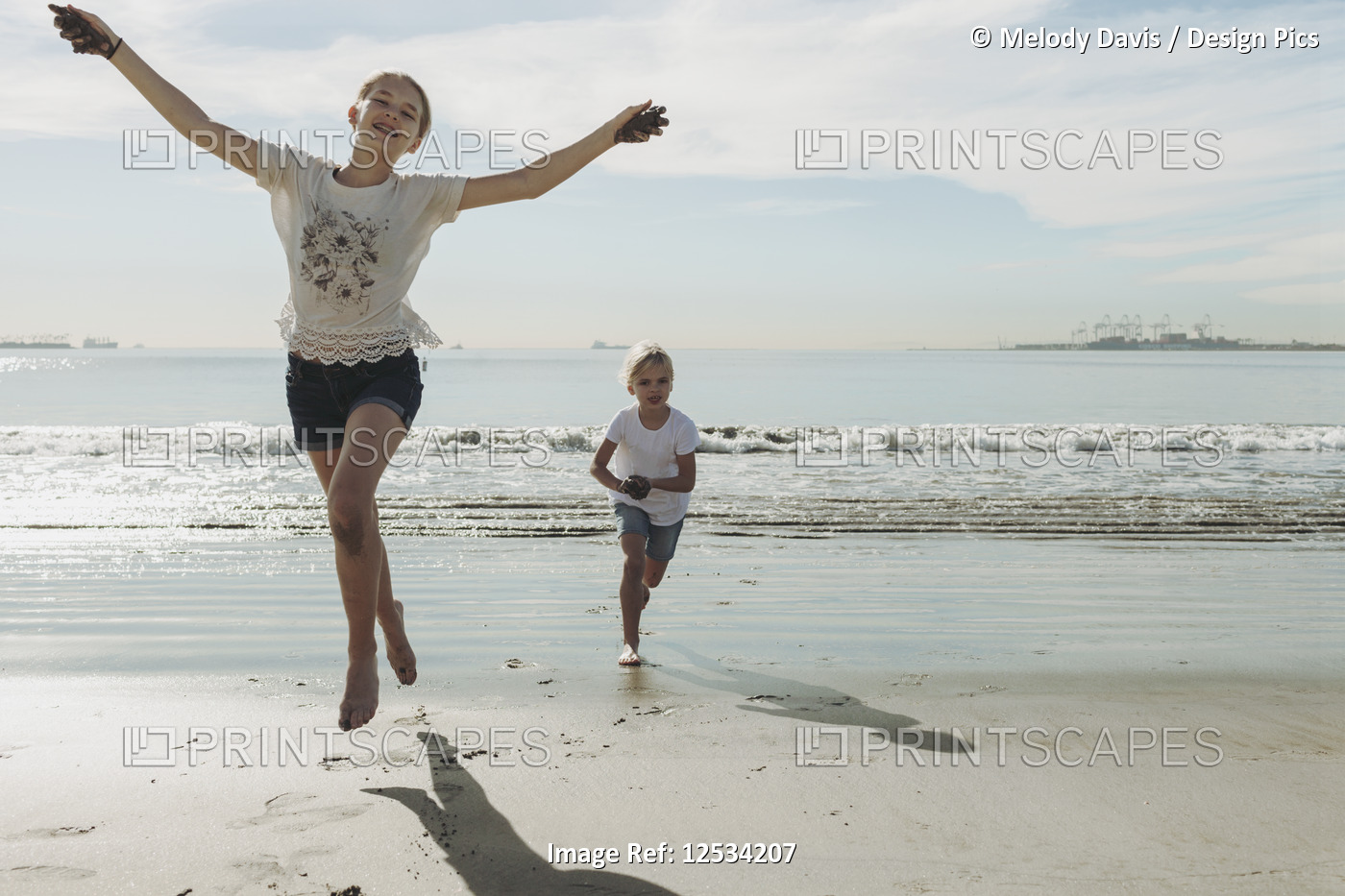 Two sisters running and playing on a beach, the oldest one in front making eye ...