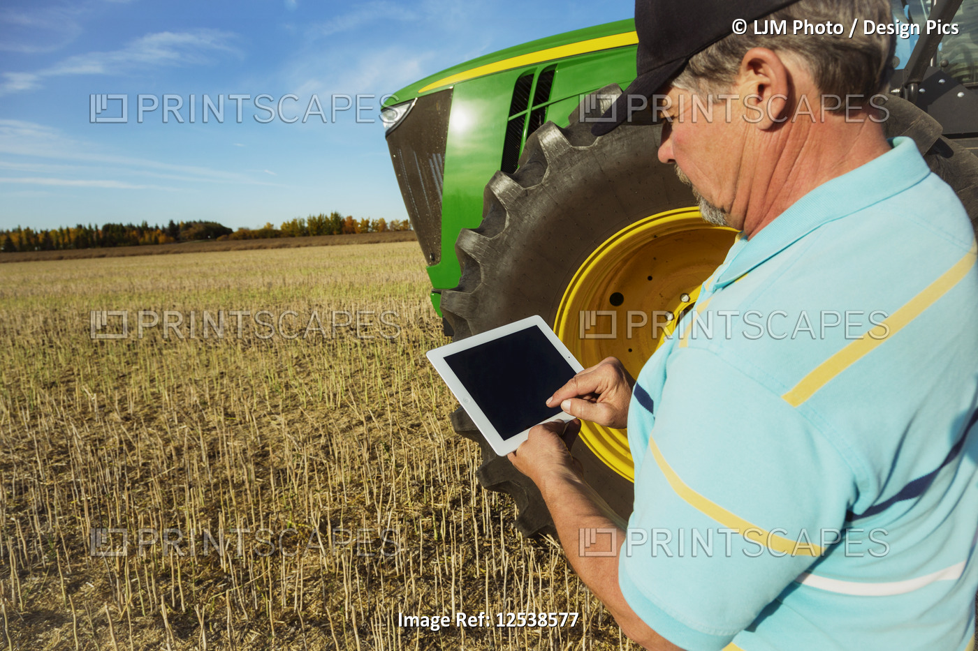 Farmer standing in a canola field beside a tractor using a tablet to assess his ...