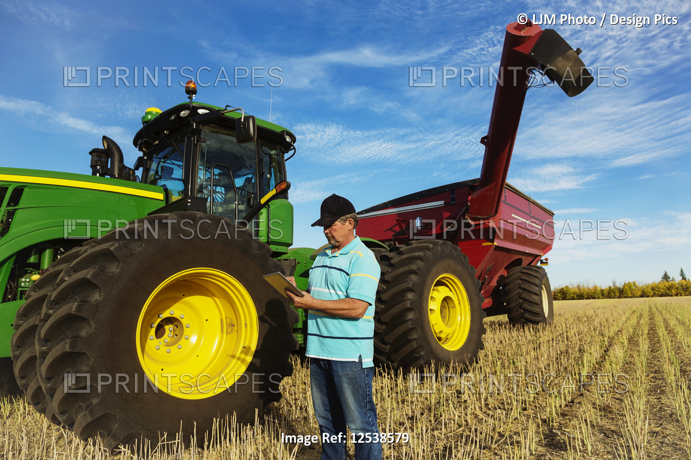 Farmer standing in a canola field beside a tractor with a wagon and using a ...