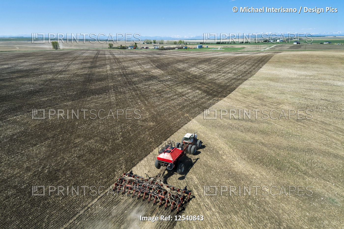 Tractor pulling an air seeder, seeding a field with mountains and blue sky in ...