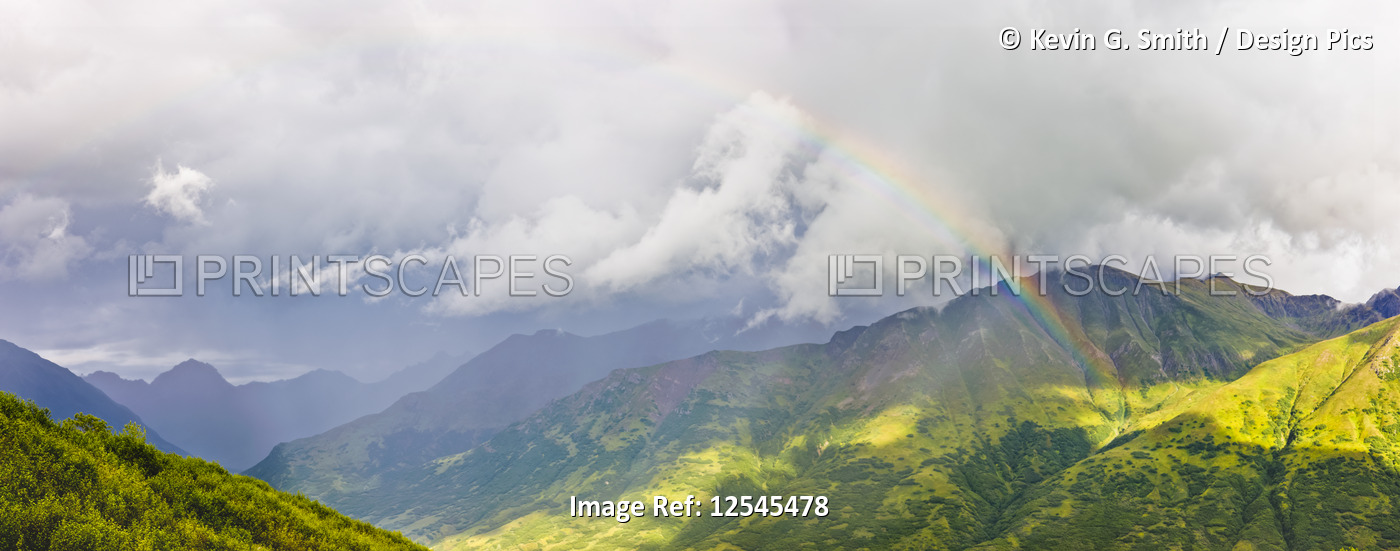 A rainbow shines through atmospheric light, lush green mountainsides in the ...