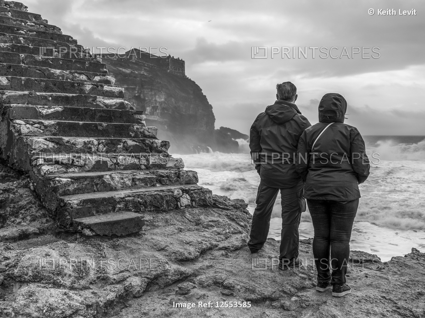 A couple stands at the water's edge looking out to the rough sea from Nazare ...
