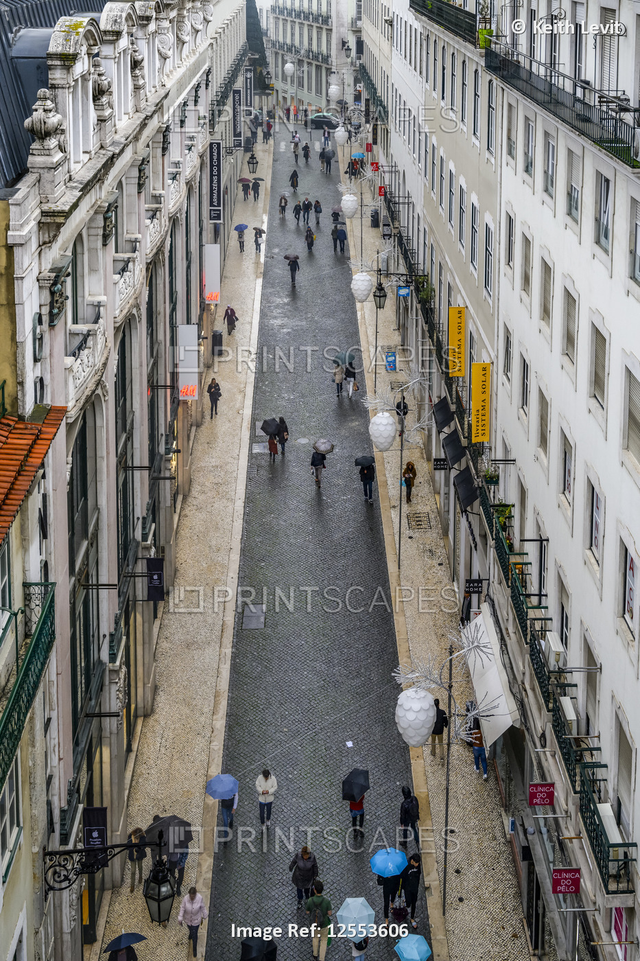 Pedestrians with umbrellas walk the narrow street in between buildings; Lisbon, ...