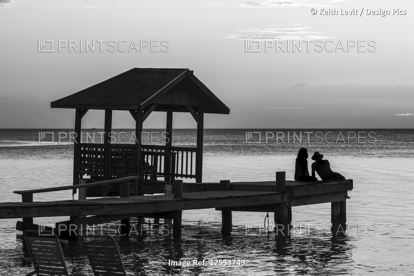 A couple sits on the end of a dock looking out to the tranquil ocean at sunset; ...