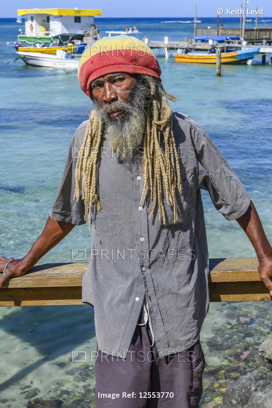 A man stands along the water's edge posing for the camera with boats moored at ...