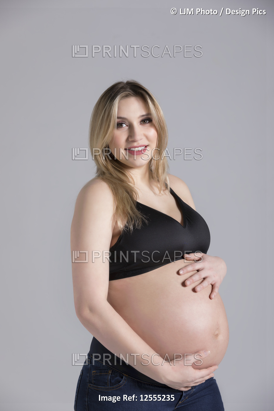 A young pregnant woman holding her belly in a studio and posing for the camera; ...