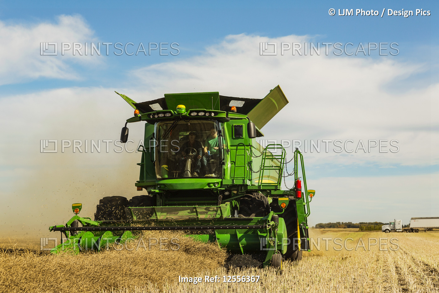 A father and son driving a combine in a field while a grain truck waits for its ...