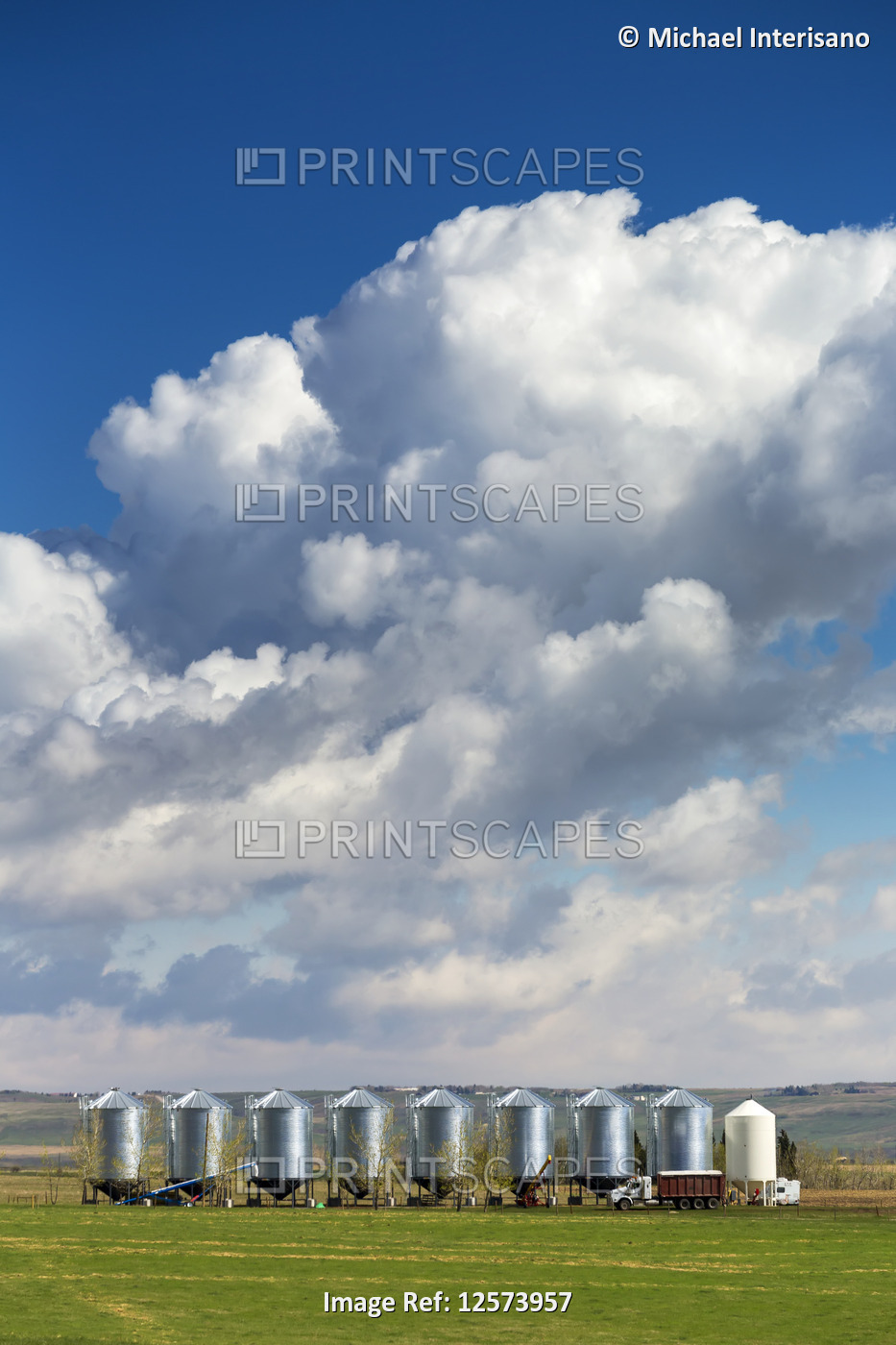 A row of large metal grain bins with dramatic storm clouds and blue sky in the ...