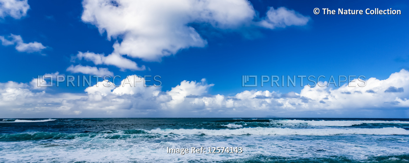 Ocean surf rolls into shore with the horizon meeting cloud and blue sky; ...