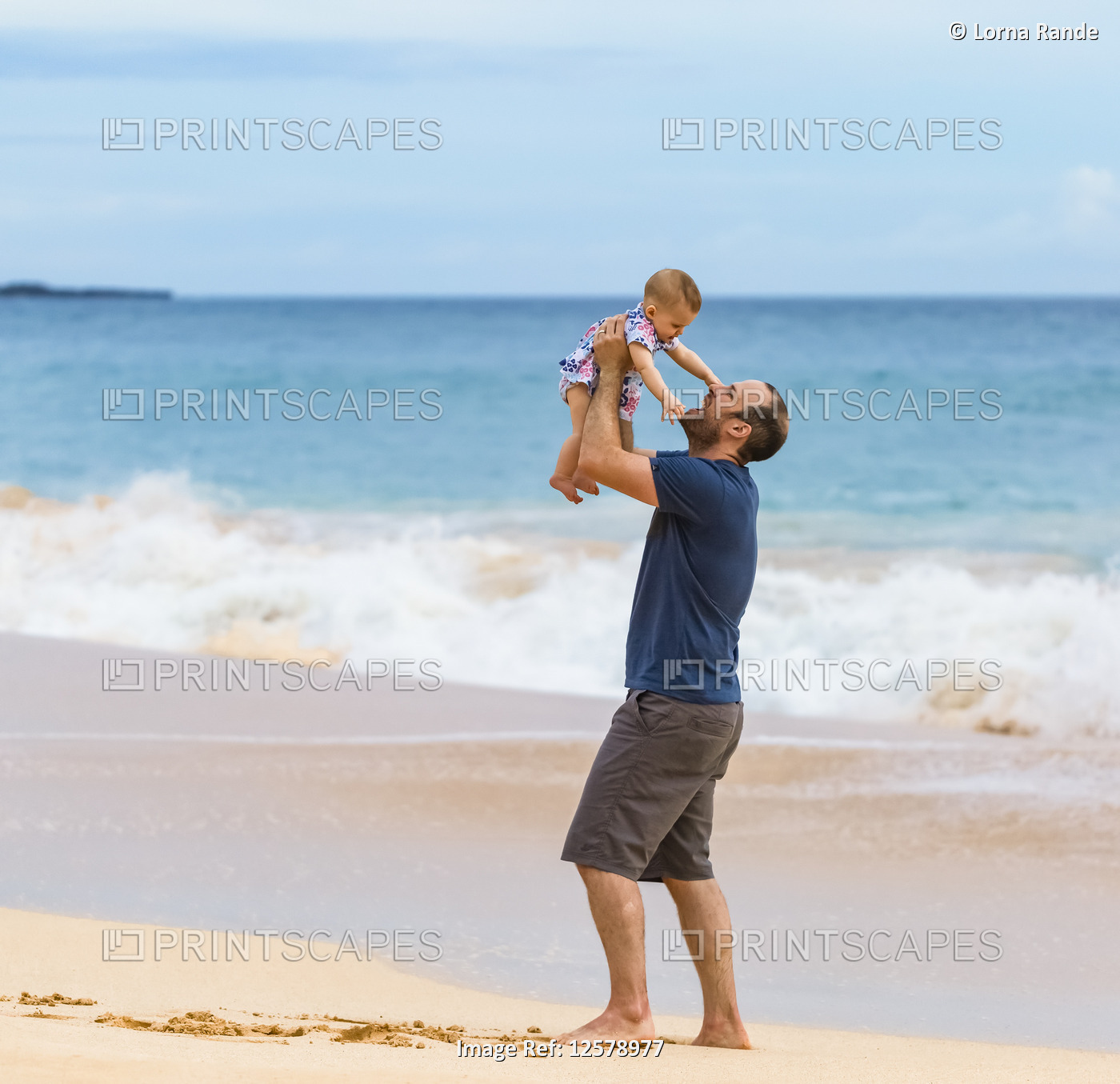 Father holds his baby daughter in the air as he stands on the beach next to the ...