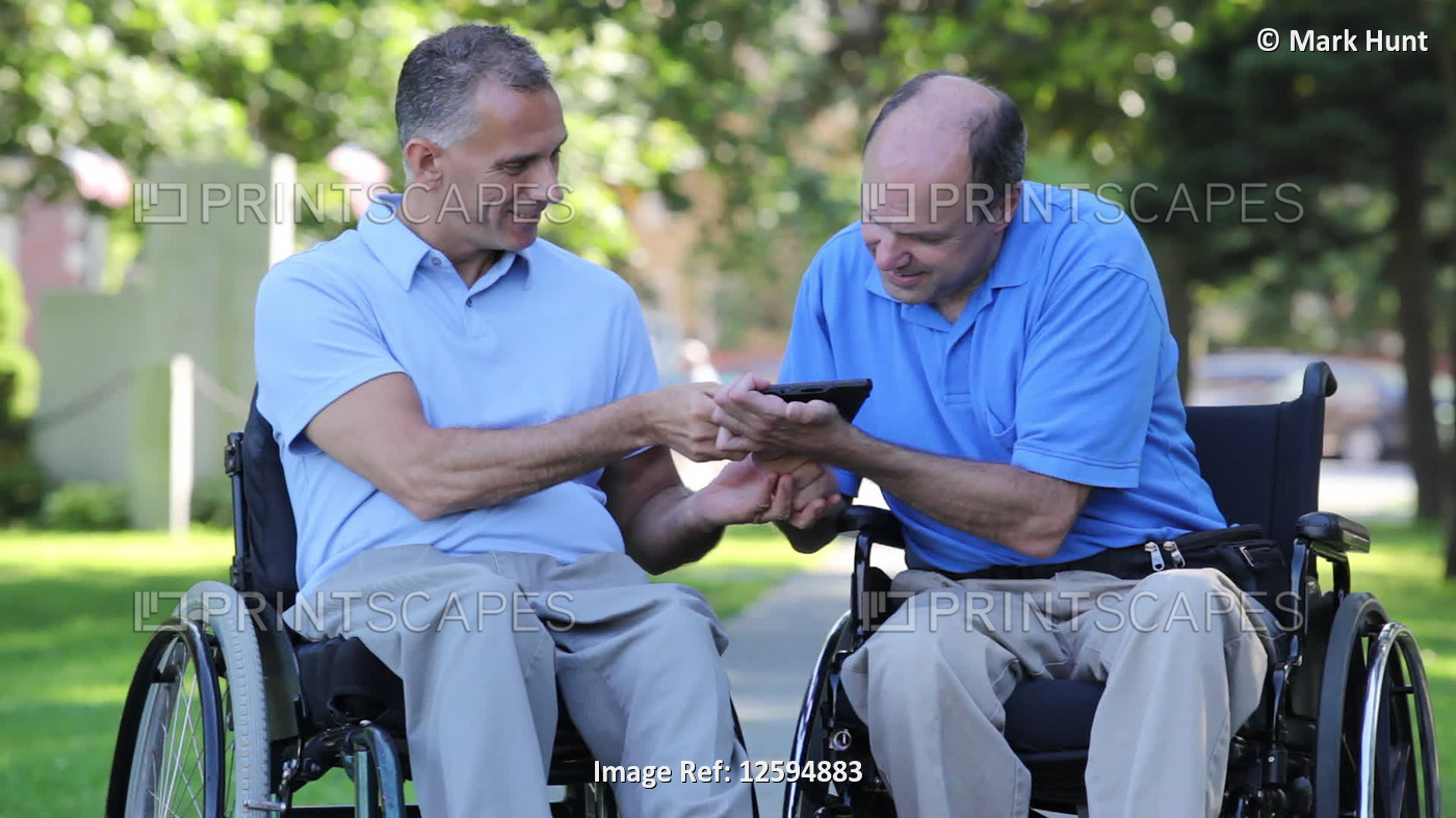 Two men in wheelchairs in a park reading a digital tablet, one with spinal cord ...