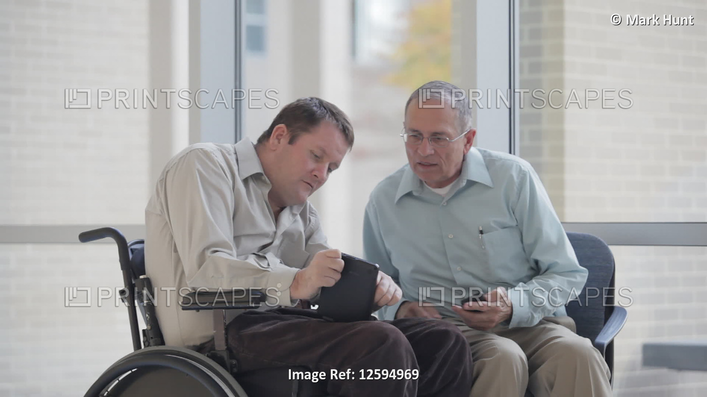 Two men comparing a tablet and a smart phone, one man with spinal cord injury ...