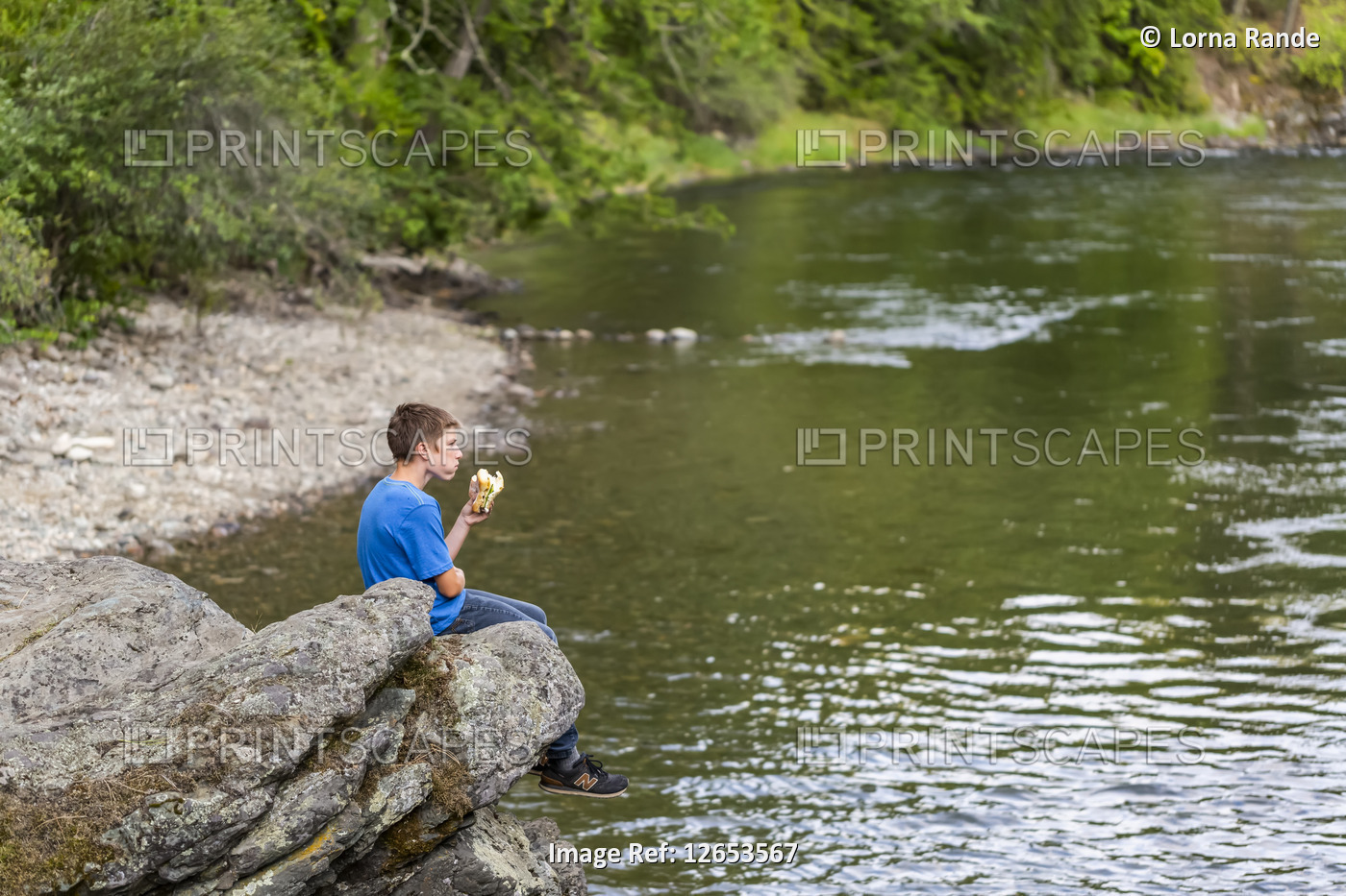 A teenage boy sits on a rock ledge over a river eating a sandwich for lunch; ...