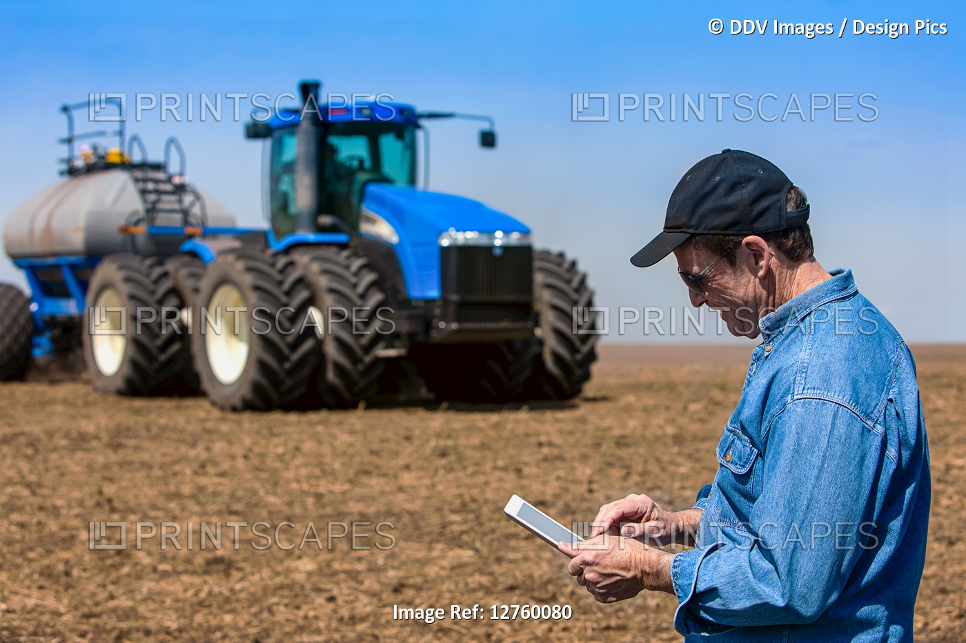 Farmer using a tablet while standing on a farm field and a tractor and ...