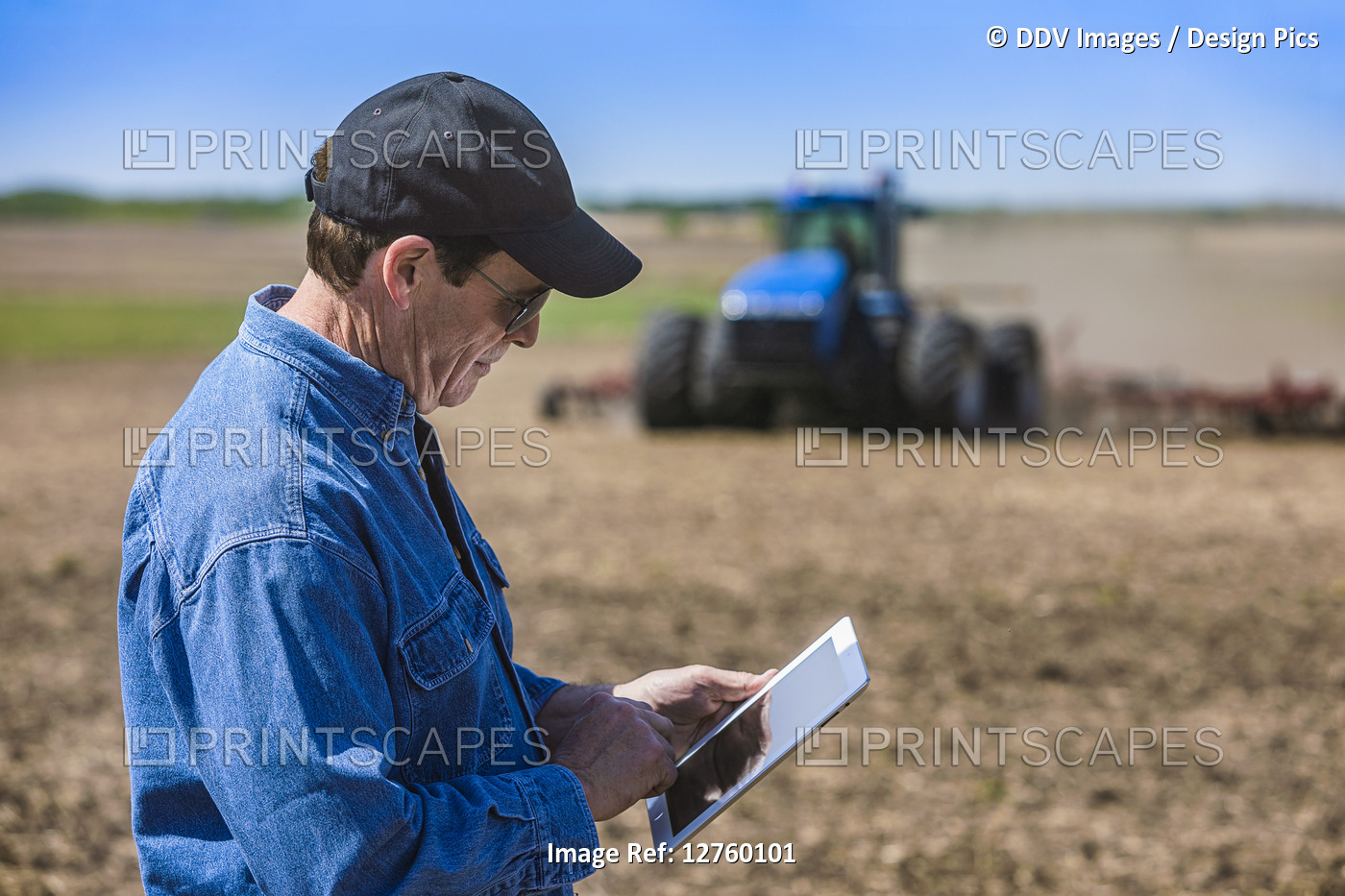 Farmer using a tablet while standing on a farm field and a tractor and ...