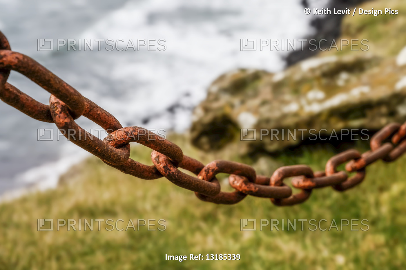 Rusted chain hanging with a coastal landscape in the background; Hafnarfjorour, ...
