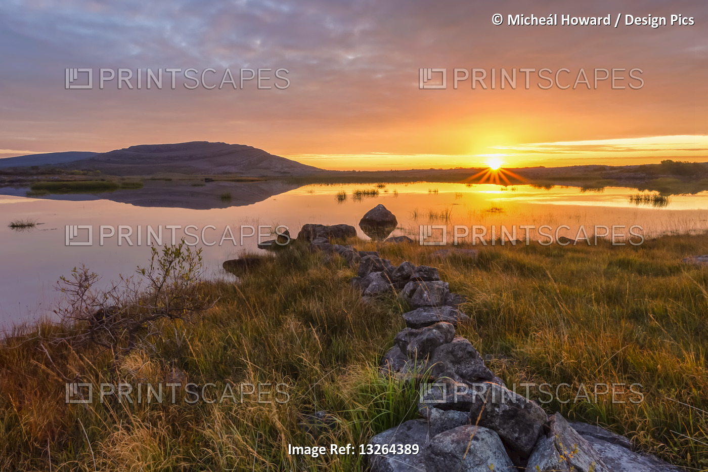 Small stone wall leading to a lake with a mountain and sunrise reflected in the ...